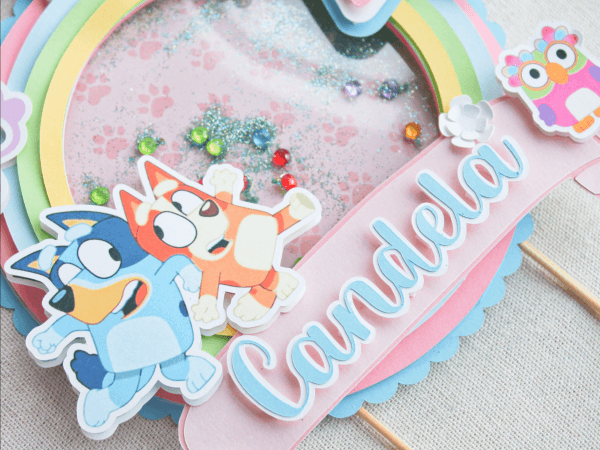 Topper para tarta personalizado, en 3D y temática Bluey. Colores Rosa, Azul y Blanco. Con motivos de los personajes, Nombre y años. Detalle Nombre, Bluey y Bingo.